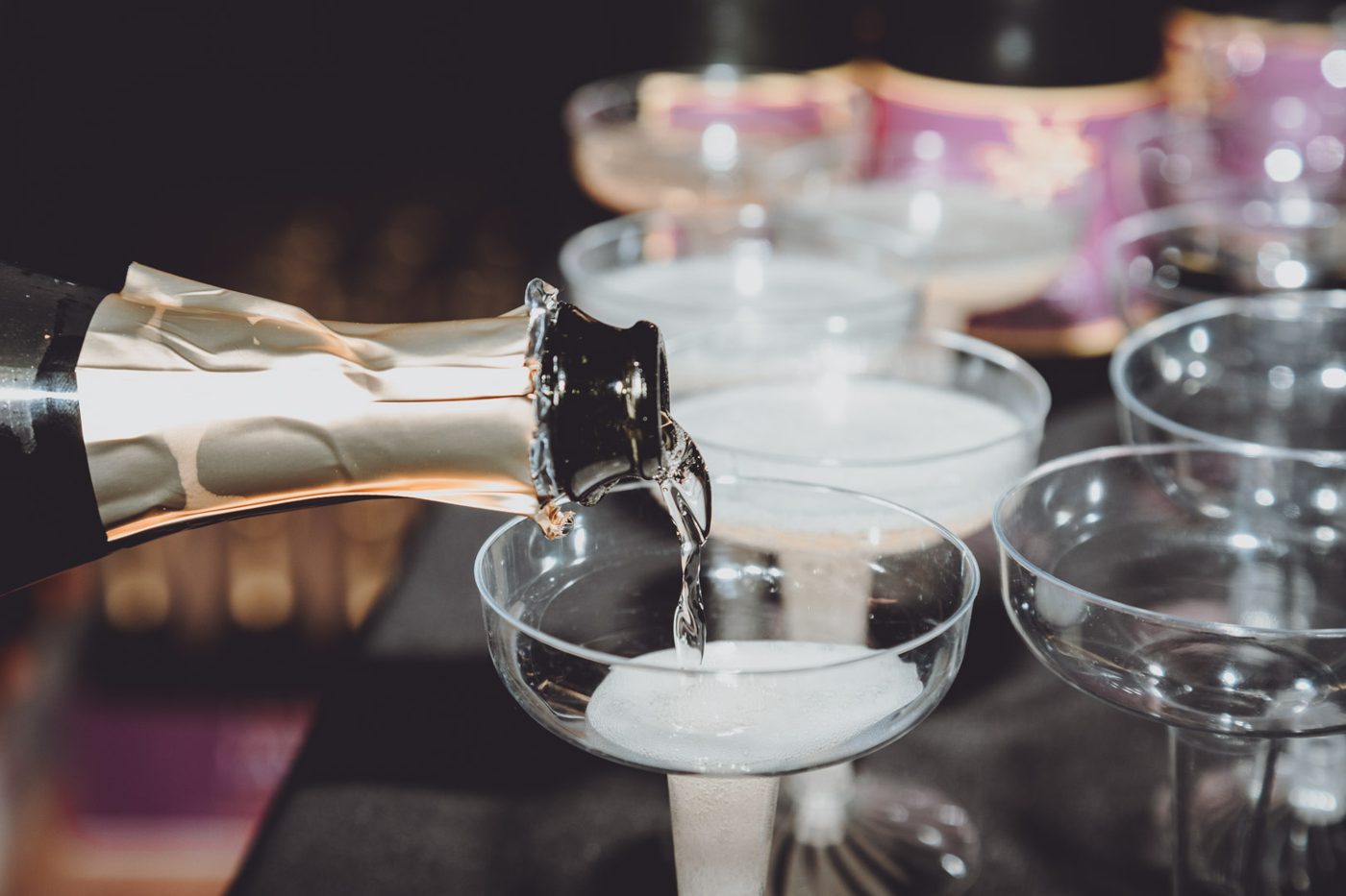 Champagne being poured into a coupe glass with more glasses and bottles behind it