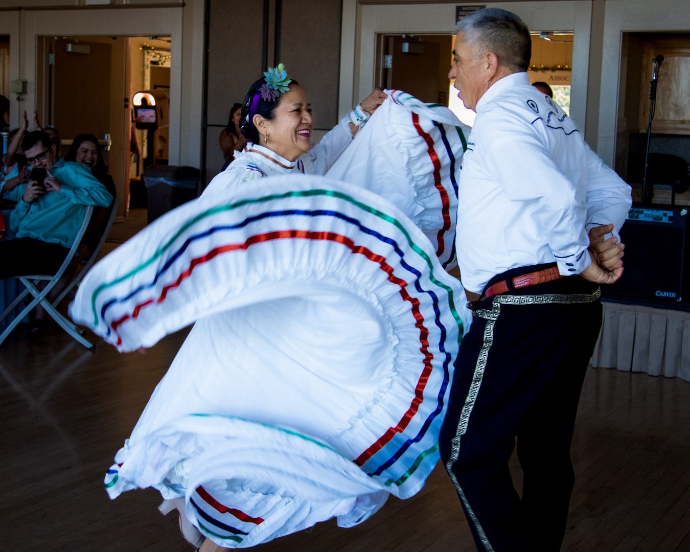 Couple in white folk costume dancing while the ribbon-trimmed skirt swirls and guests laugh behind them