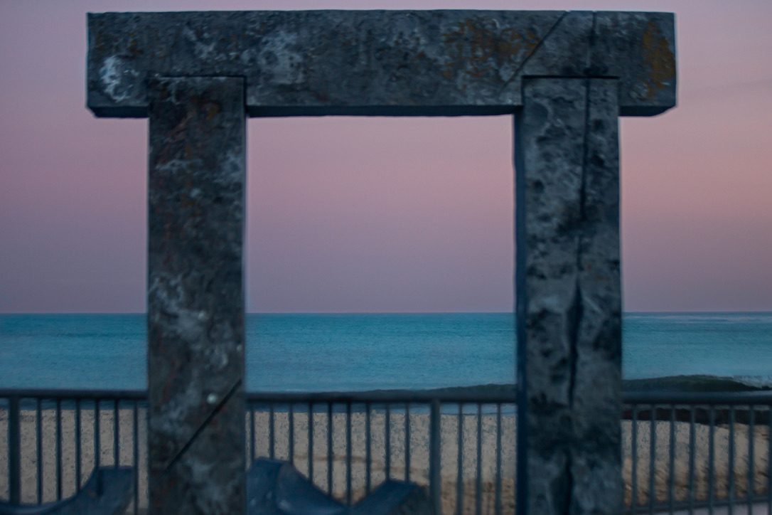 Weathered stone archway framing a rose and lavender dusk sky over the ocean, with a railing and beach below