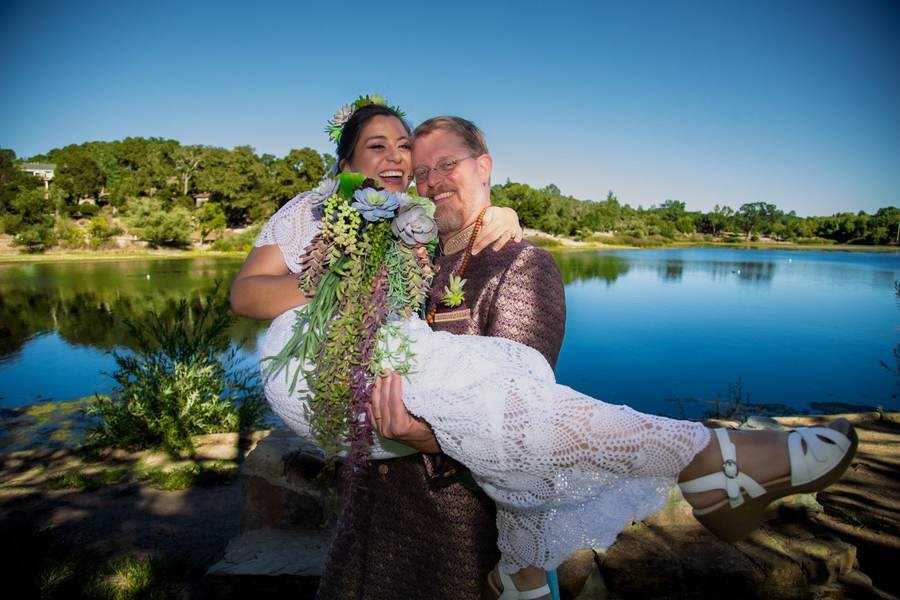 Groom carrying bride laughing by a lake in bright sunlight