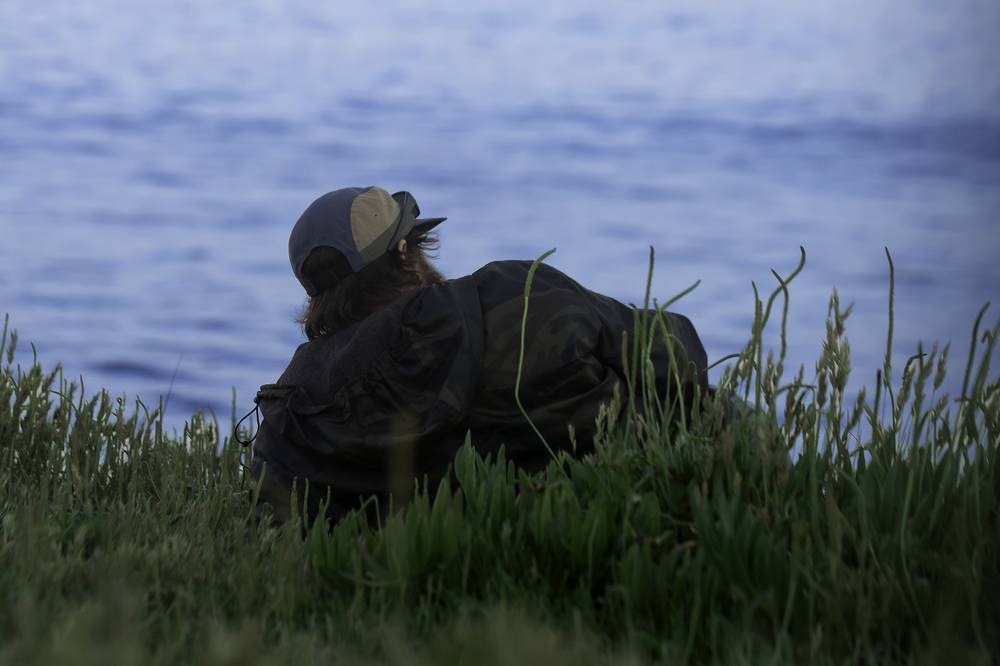 Person in a cap and camo jacket sitting alone watching the ocean at dusk