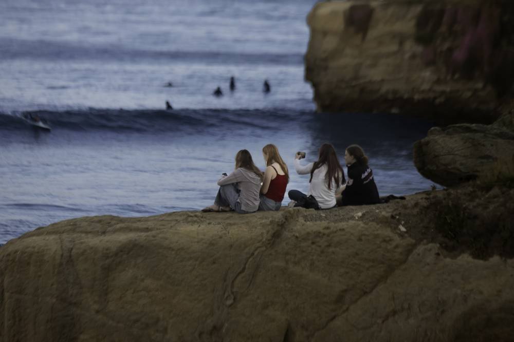 Four friends sitting on cliff rocks watching surfers at dusk