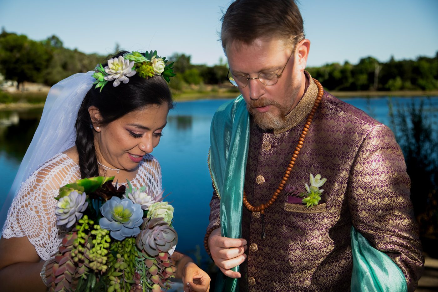 Bride in a succulent crown and groom in a purple and gold jacket looking down together beside a lake