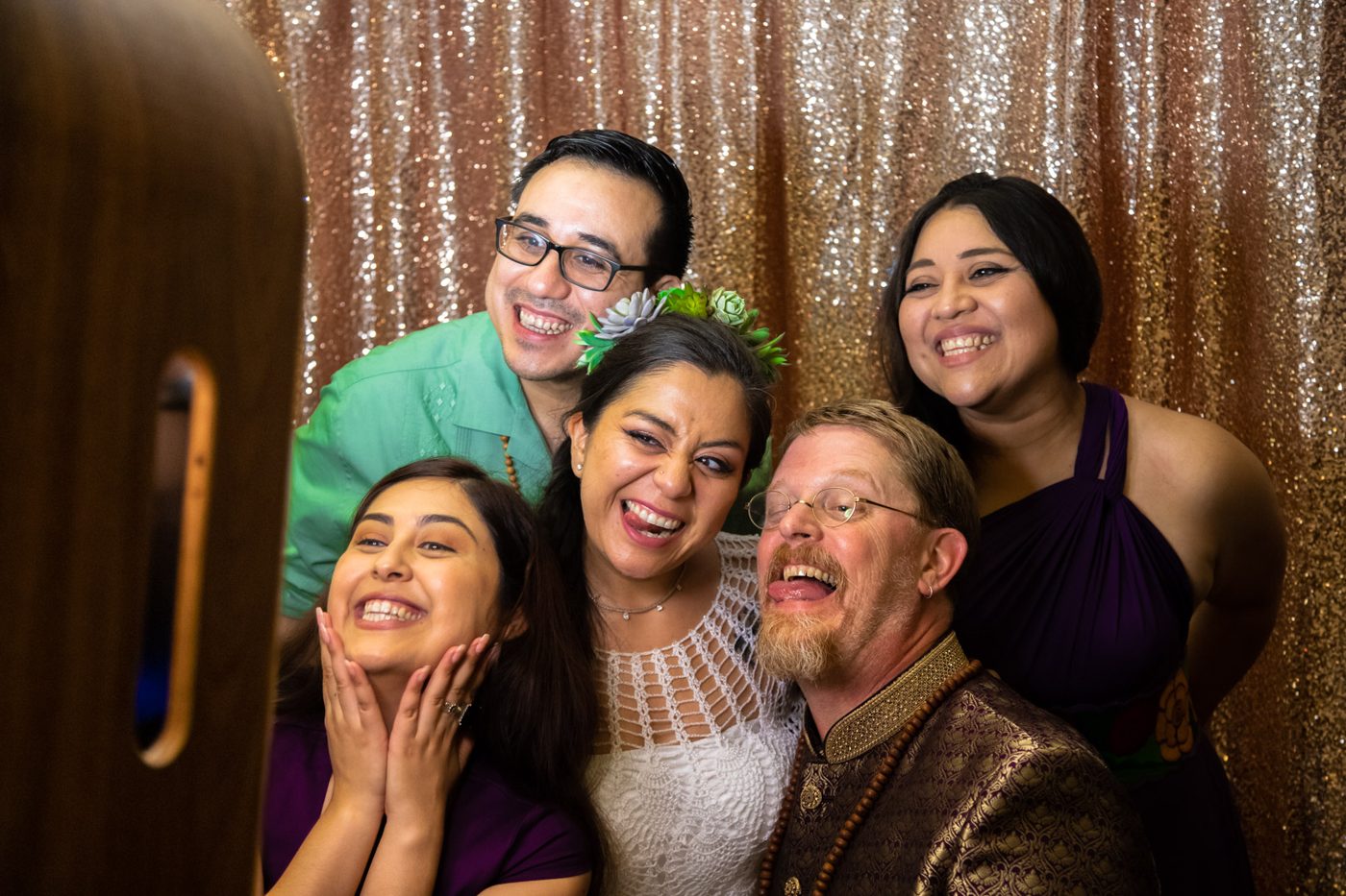 Bride, groom and three friends laughing and making faces in front of a sequin photo booth backdrop