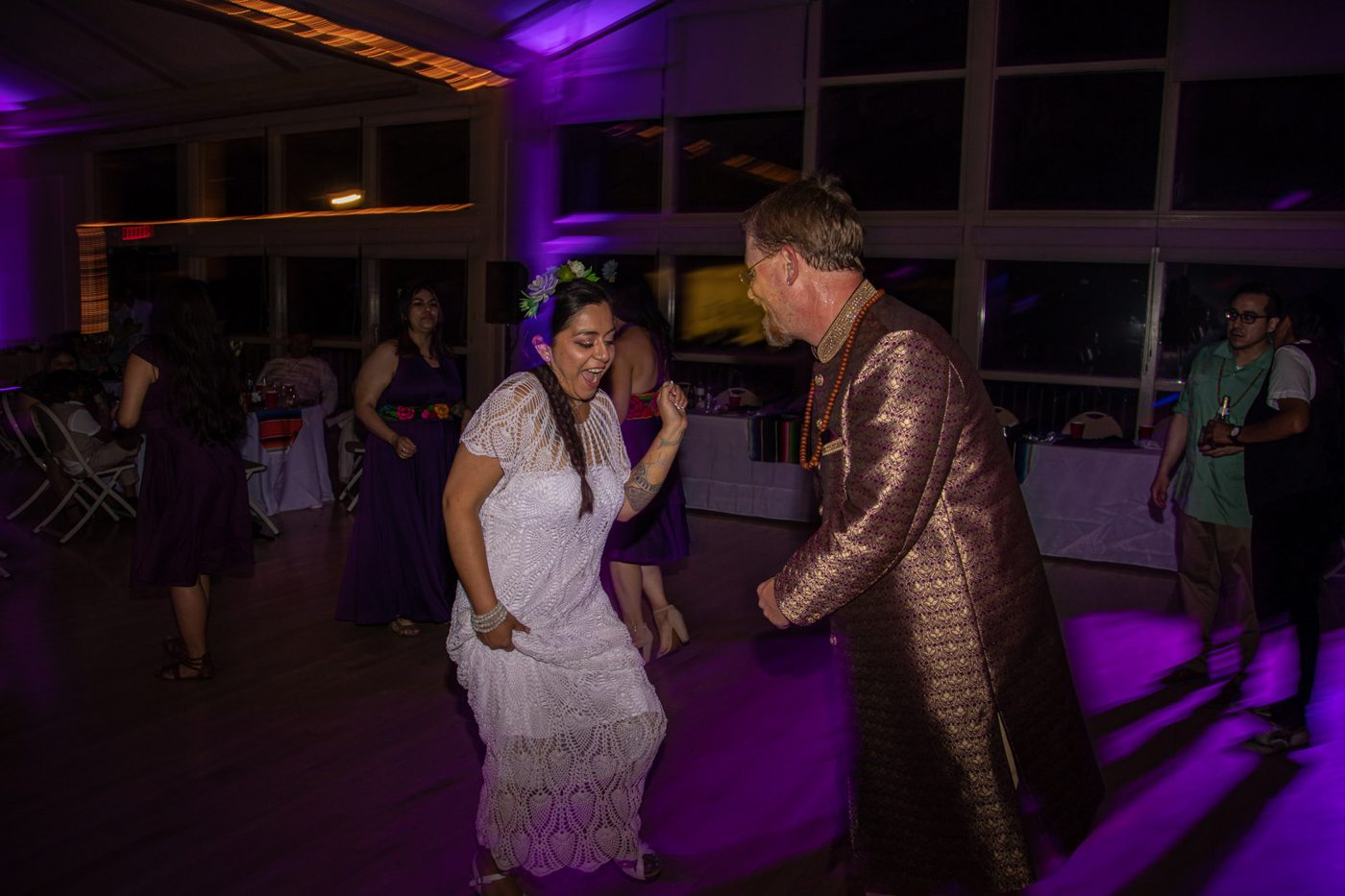 Bride lifting the hem of her crocheted dress while dancing with the groom under purple light