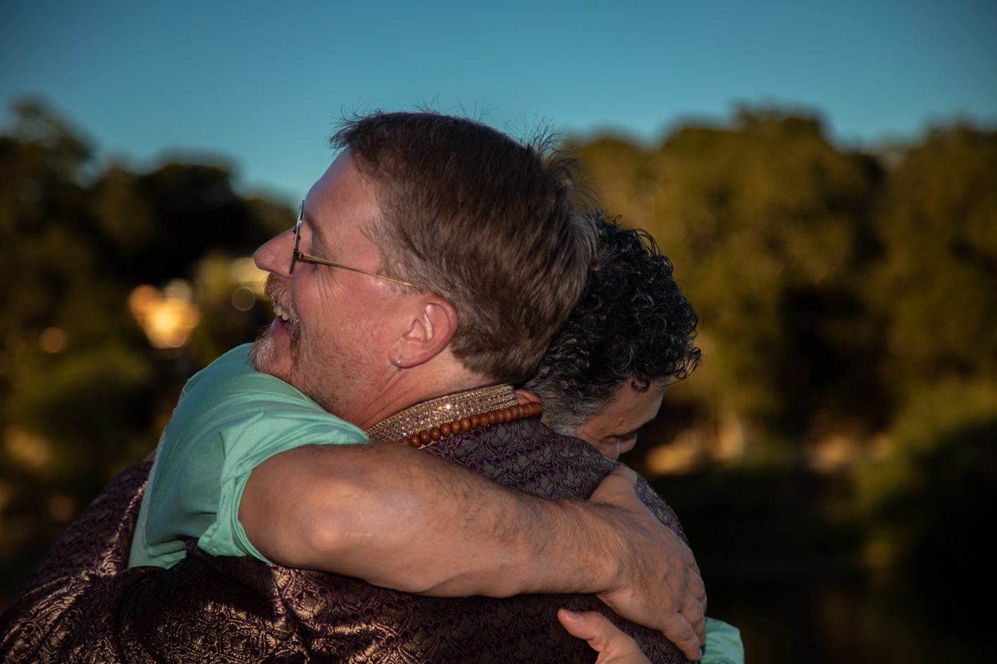 Groom in a purple and gold jacket laughing as he hugs a guest at golden hour