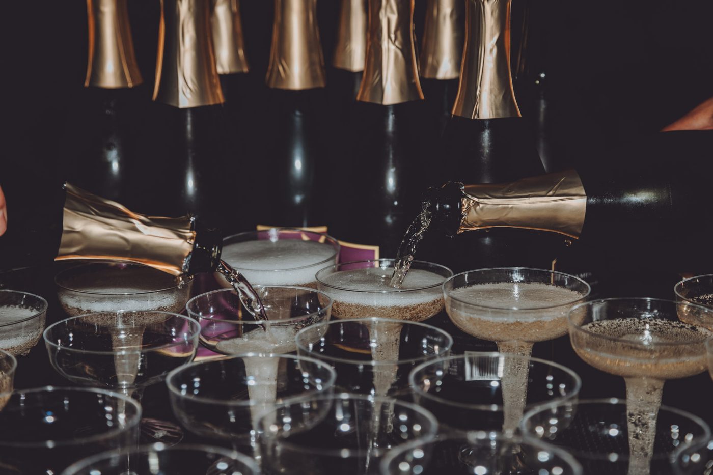 Two bottles pouring champagne into rows of coupe glasses against a dark background