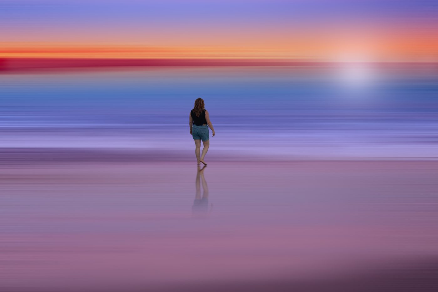 Woman walking barefoot on a wet beach into a long-exposure sunset of blurred orange, blue and violet
