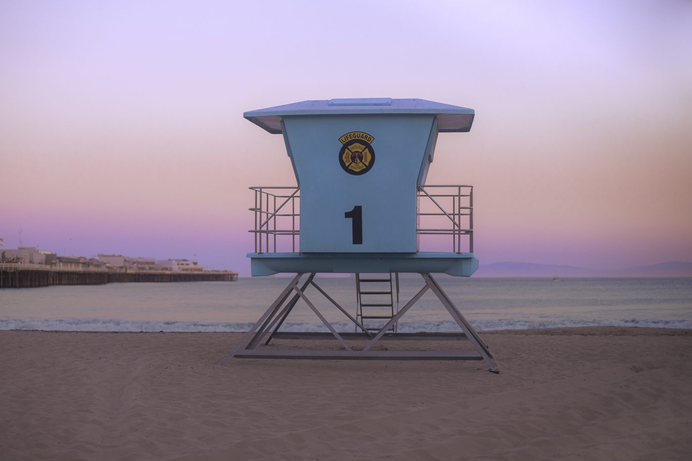 Blue lifeguard tower number 1 on the sand with the wharf and calm ocean under a pink and lavender sky